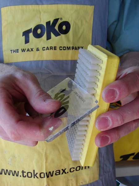 Clean brush by aggressively rubbing a scrapper edge against it, going back and forth